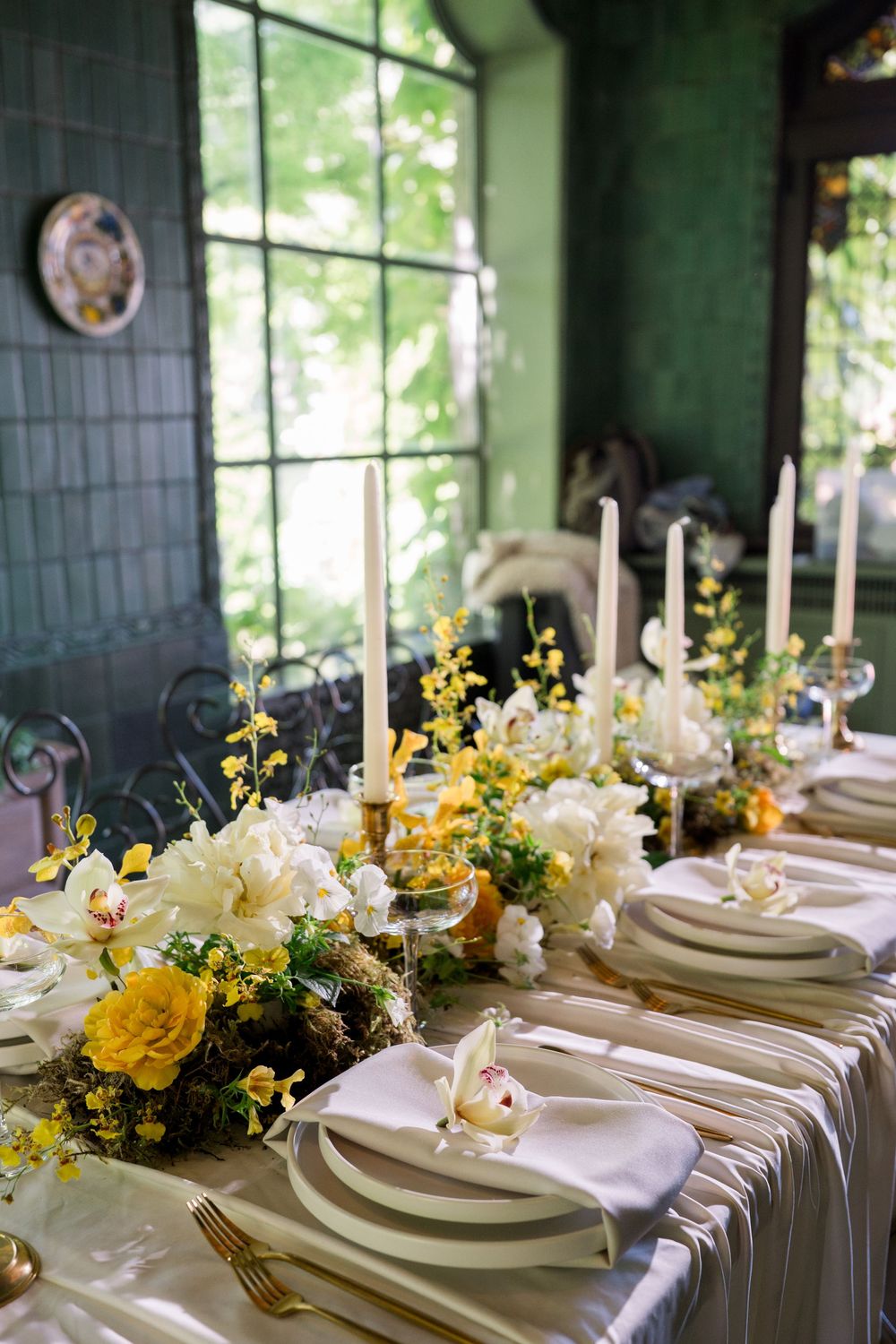 Elegant wedding table setting with white linens, gold cutlery, white candles, and yellow floral centerpieces in a green venue.