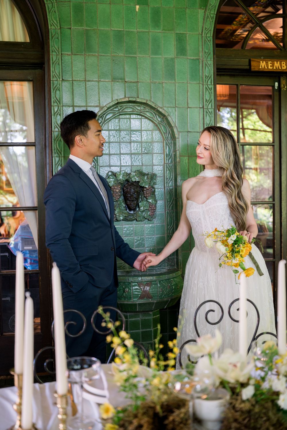 Bride and groom hold hands before green tiled wall at elegant wedding venue with floral arrangements.