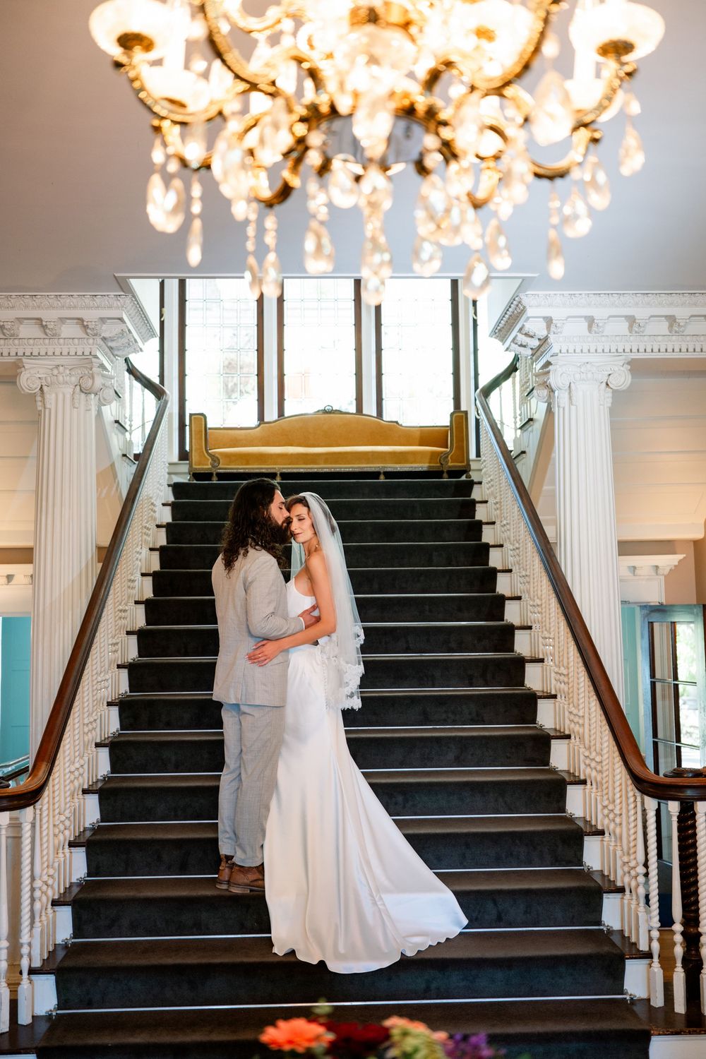 Bride and groom share romantic moment on grand staircase beneath crystal chandelier in elegant wedding venue.