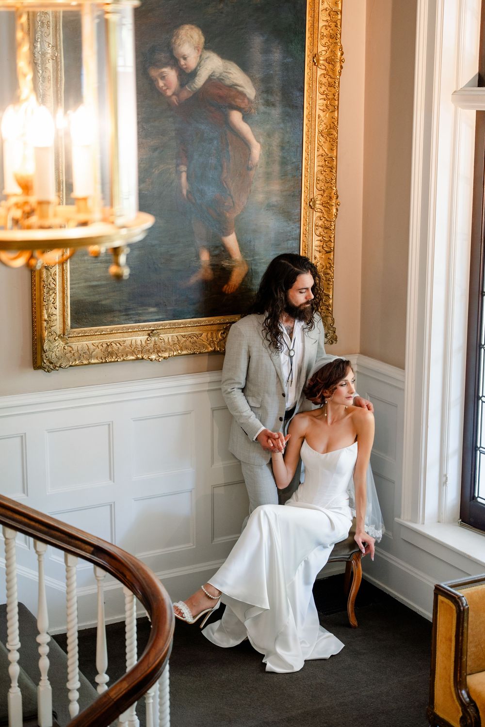 Bride in white gown seated by window with groom in gray suit beside her, elegant room with gold-framed painting.
