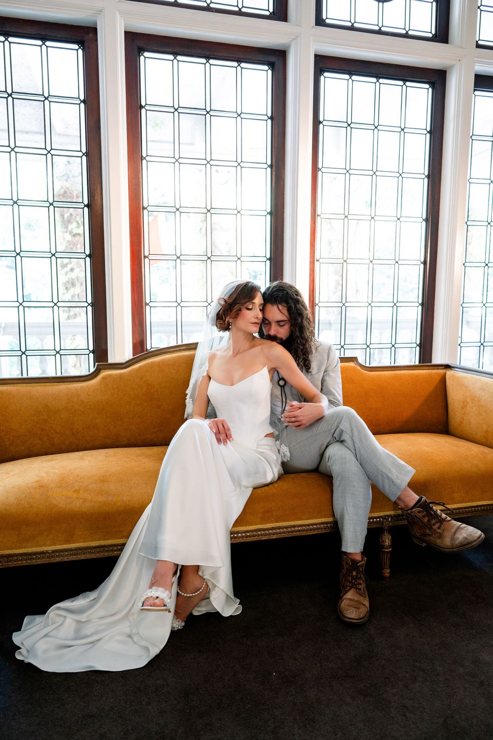 Bride and groom share an intimate moment on a gold velvet sofa in front of large leaded glass windows.
