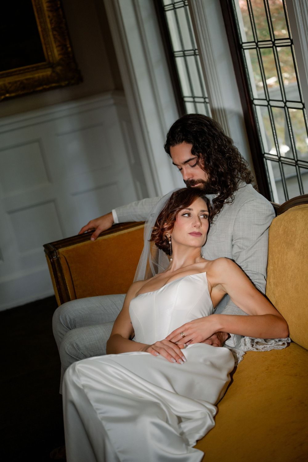 Bride in white gown rests against groom in gray suit on a gold velvet chair near leaded glass windows.