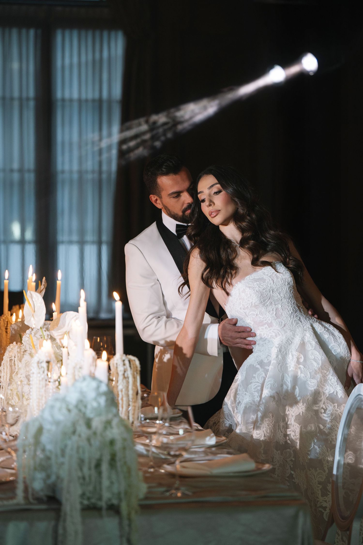Bride and groom in elegant attire seated at candlelit reception table in dark venue.