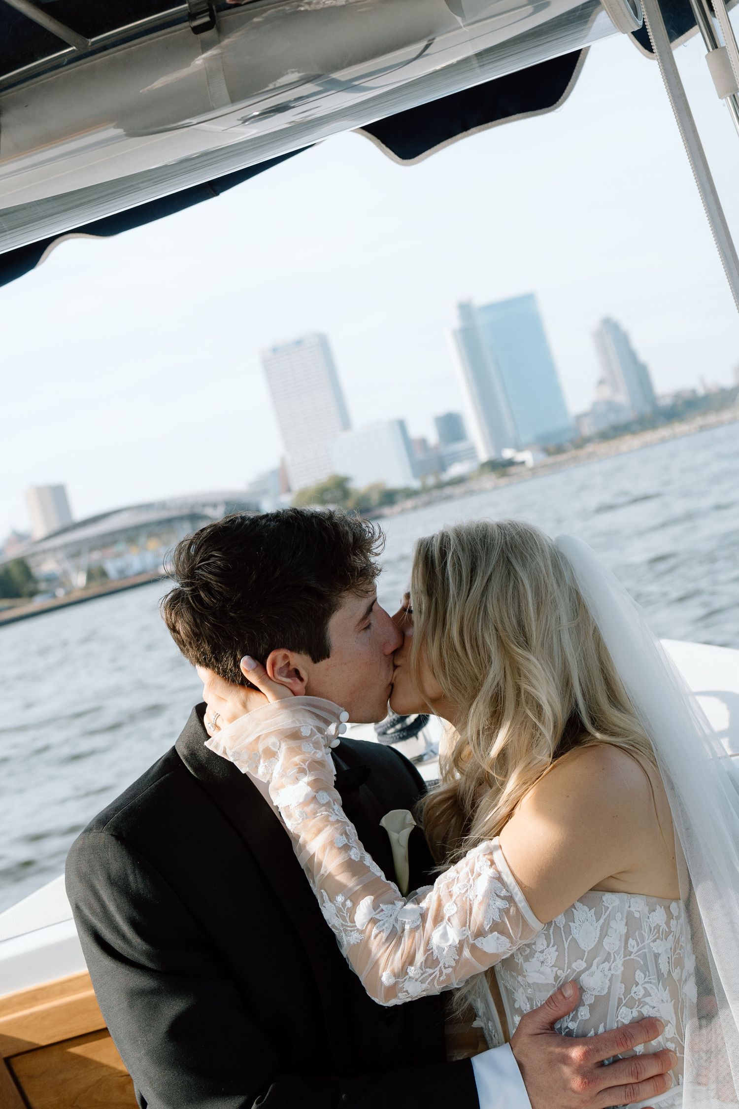 A romantic couple shares a tender kiss while sitting on a boat with a city skyline visible in the background.