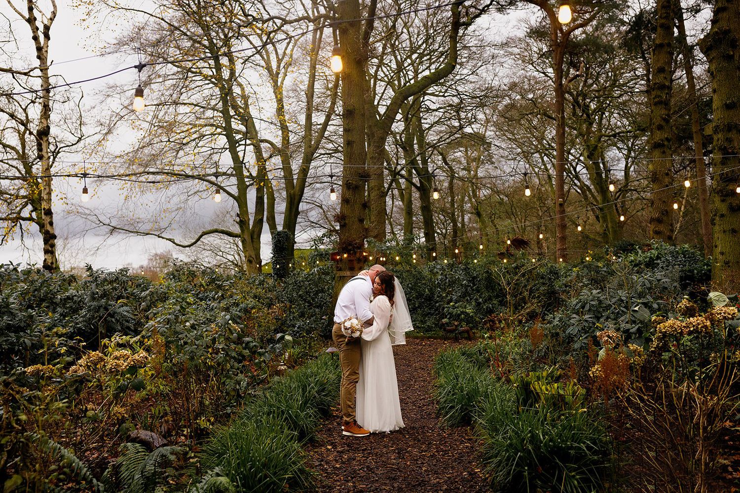 A couple in white attire embraces among autumn trees during a romantic outdoor wedding photoshoot.