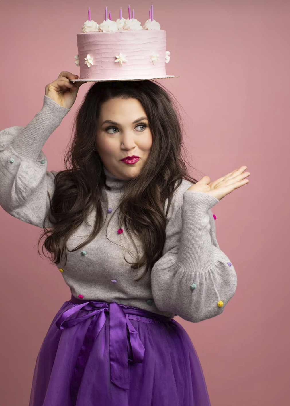 A model in a gray sweater and purple skirt poses playfully with a pink birthday cake crown against a pink backdrop. Elizabeth Lang Photograp