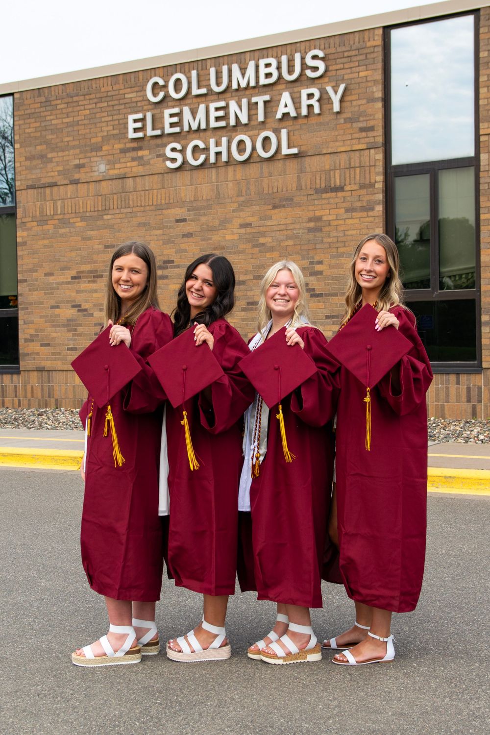 A group of graduates in maroon gowns pose in front of Columbus Elementary School.