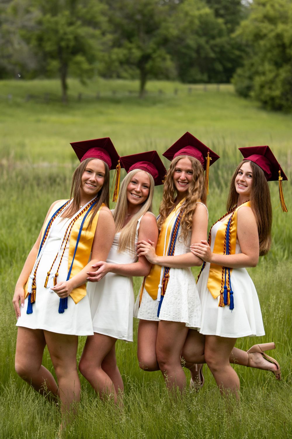Graduates in white dresses and maroon caps pose playfully in a sunny meadow.