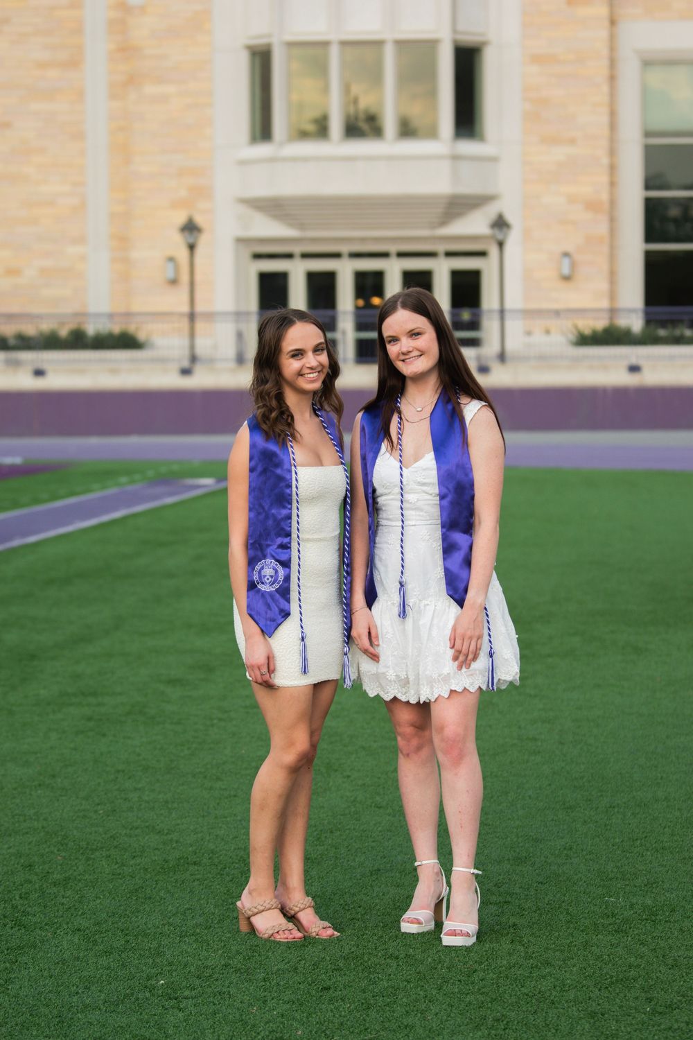 Two graduates in white dresses and blue sashes pose on a college campus field.