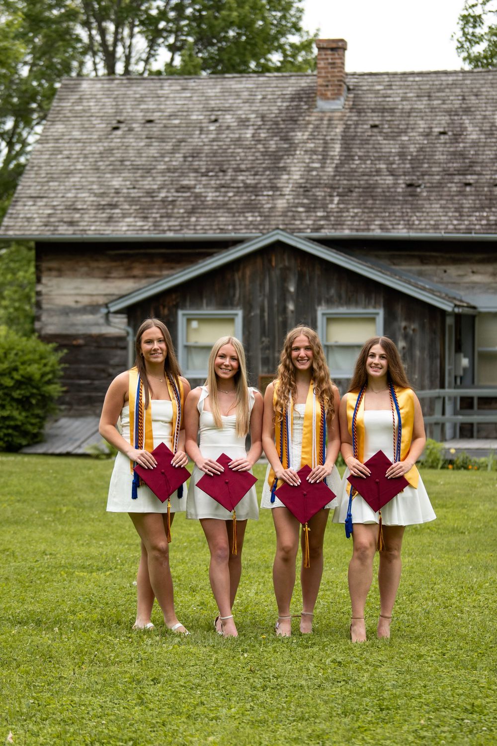 Friends in white dresses hold maroon graduation caps while posing in front of a rustic cabin.