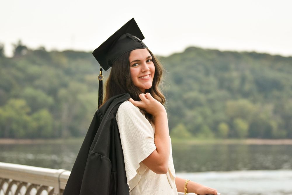 Graduate in cap and gown poses outdoors near waterfront.