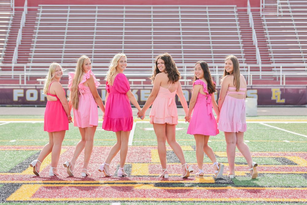 Group wearing pink dresses sits together on football field bleachers.