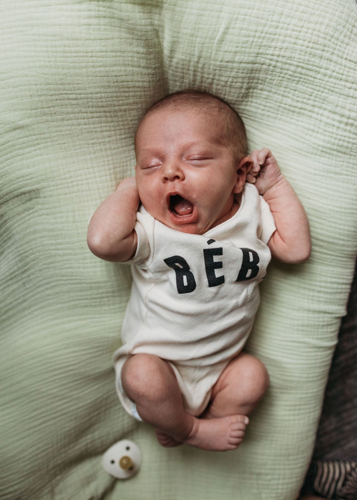 A baby lies on a light green blanket wearing a 'BE BOLD' onesie while holding a pacifier.