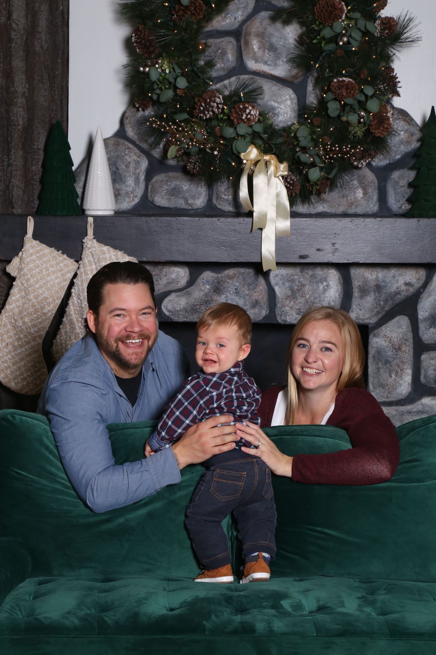 A festive holiday scene with a stone fireplace backdrop, decorated wreath, and green velvet couch.