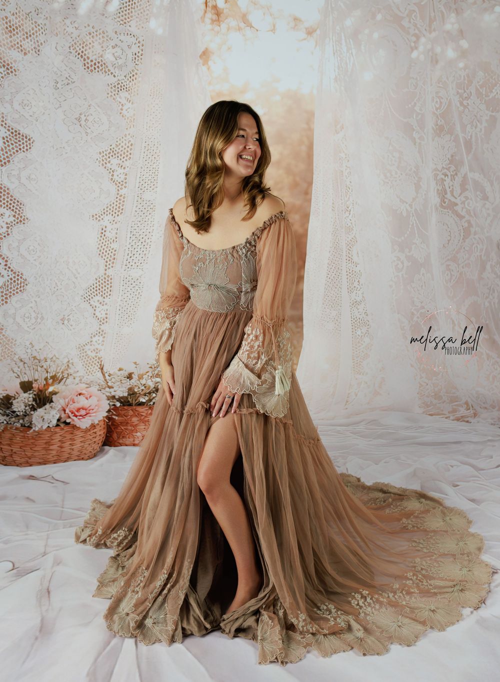 A flowing beige vintage-style dress with lace details and ruffles photographed against a light backdrop with floral accents.