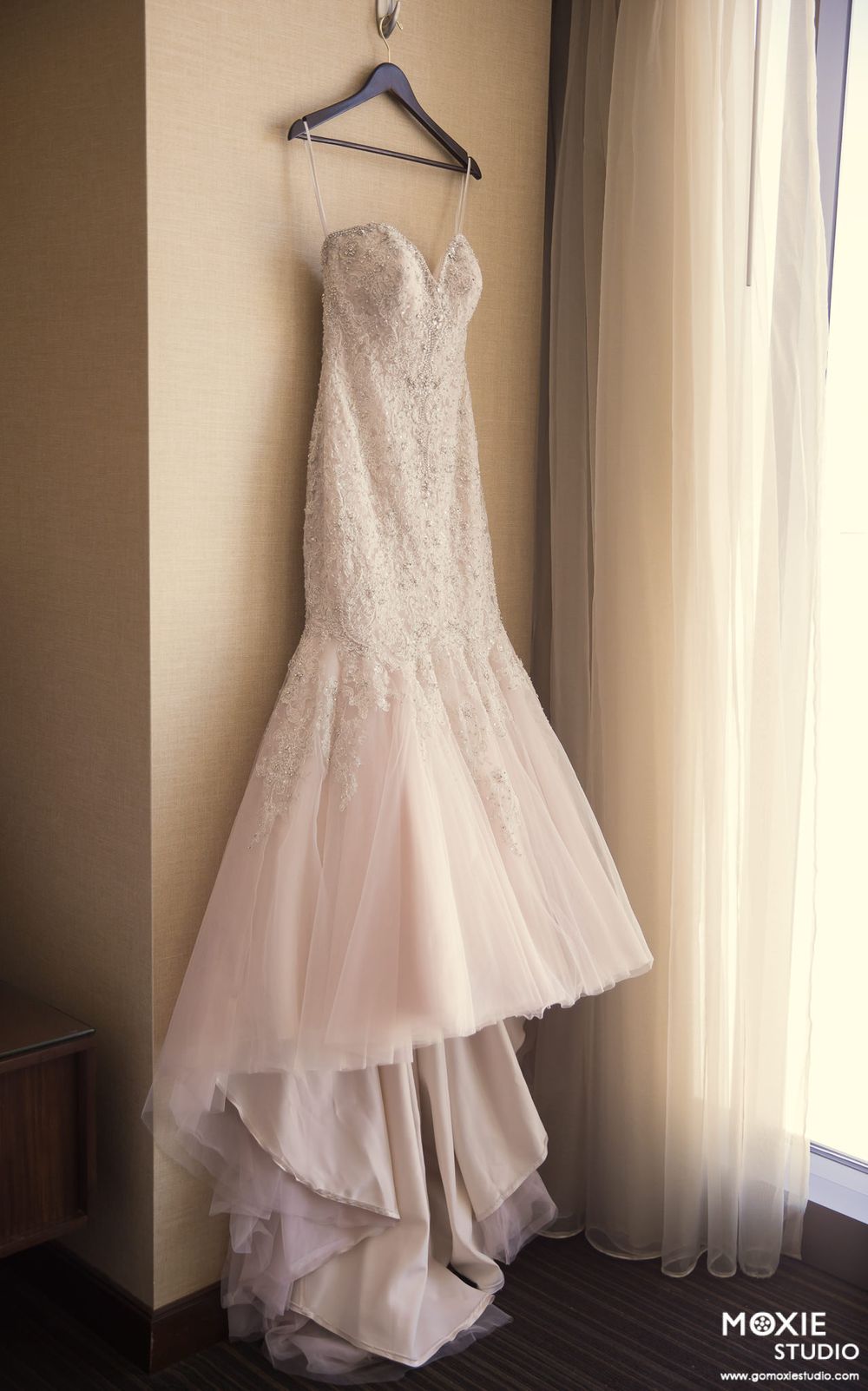 A delicate white lace mermaid wedding gown with sweetheart neckline hangs elegantly on a blue hanger against a beige wall.