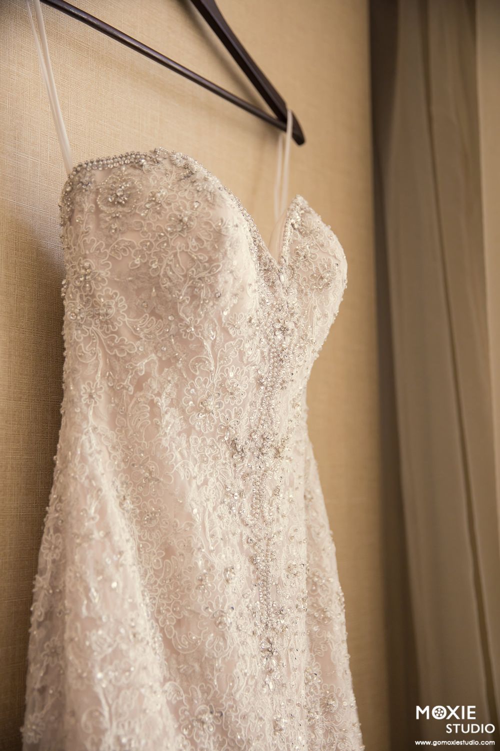 A delicate white lace wedding dress with intricate beading hangs on a black hanger against a beige wall.