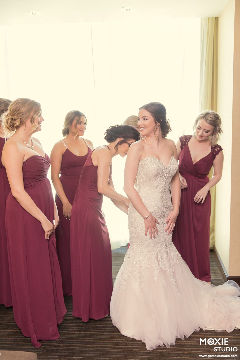 A bridal party in burgundy bridesmaid dresses and a white wedding gown share candid laughter together.