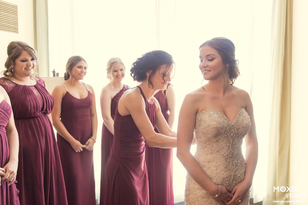 Bridesmaids in burgundy dresses help bride prepare for wedding ceremony in sunlit room.