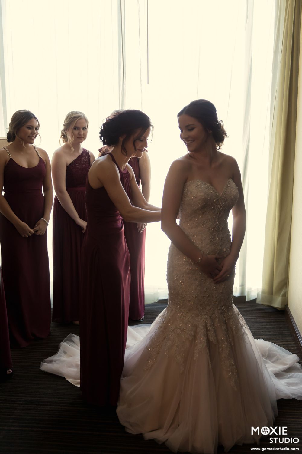 Bridesmaids in burgundy dresses help the bride with her flowing tulle wedding gown.