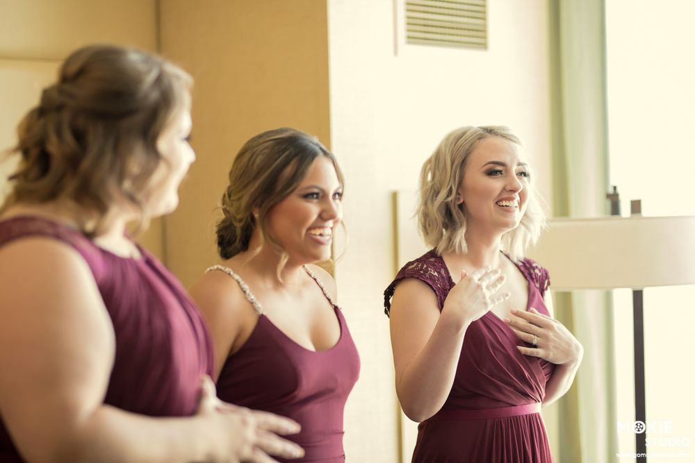 Bridesmaids in burgundy dresses share joyful laughter while getting ready in a sunlit room.