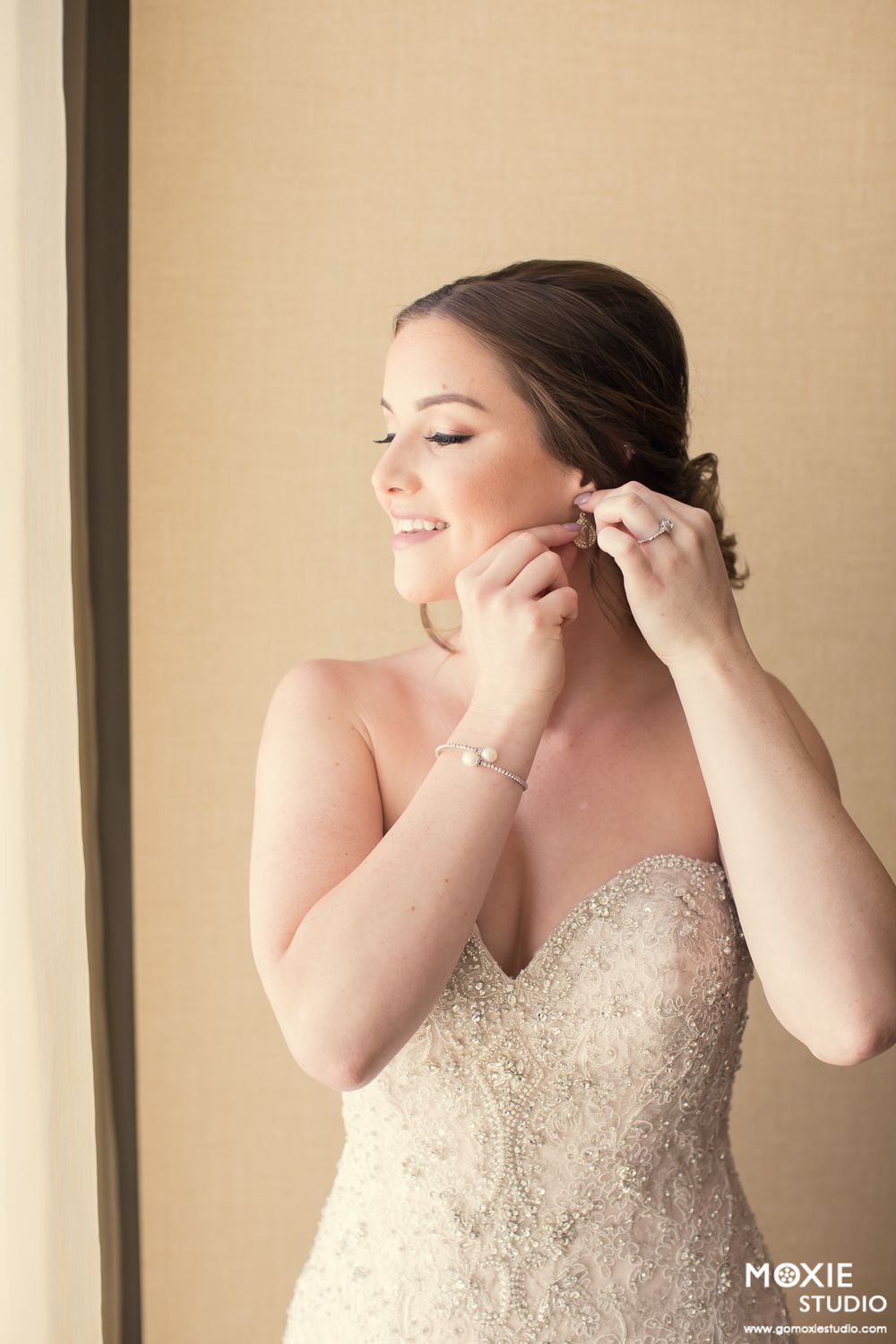A woman in a strapless beaded wedding gown puts on earrings while getting ready.