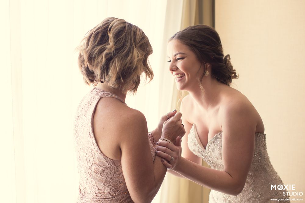 Bridesmaids in burgundy dresses help the bride with final wedding day preparations in a sunlit room.
