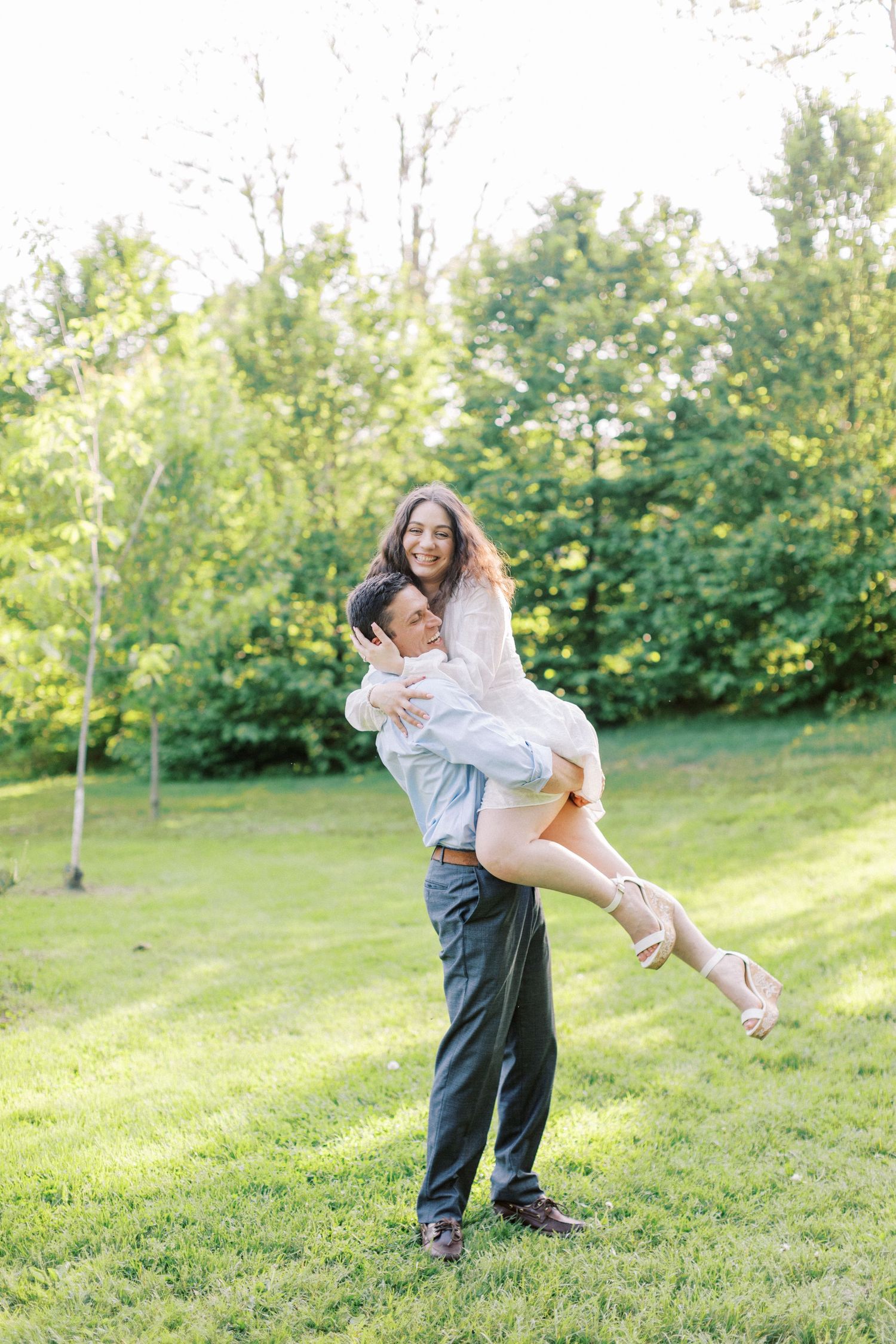 A couple embraces and spins playfully together on a grassy hillside surrounded by green trees on a sunny day.