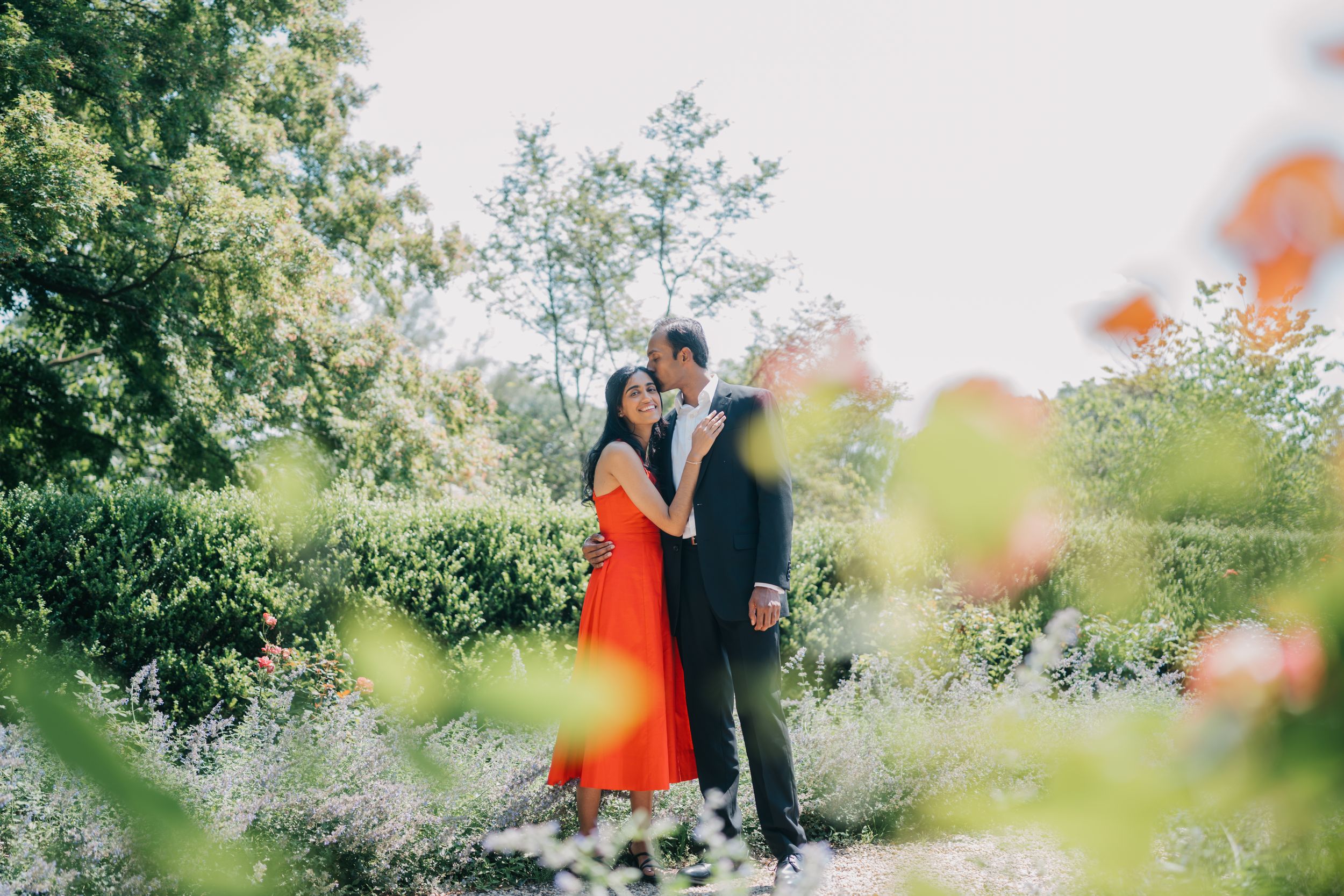 A couple stands together in a lush garden setting surrounded by blooming flowers and greenery.