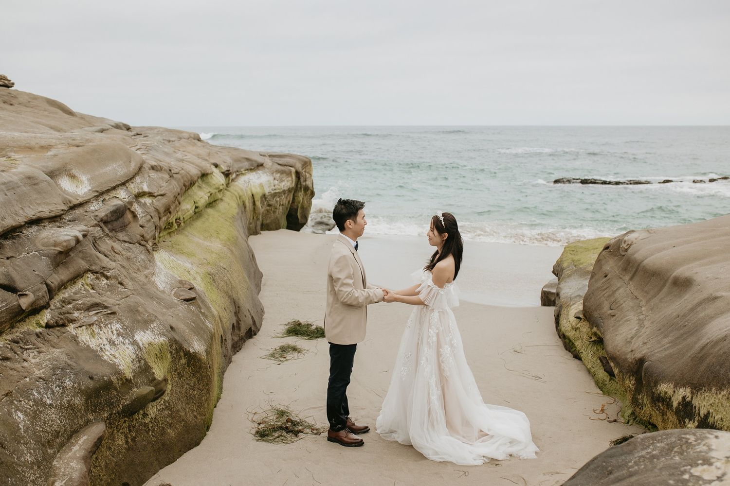 A couple stands together on a scenic beach surrounded by rocky cliffs and turquoise ocean waters at Windansea Beach in La Jolla.