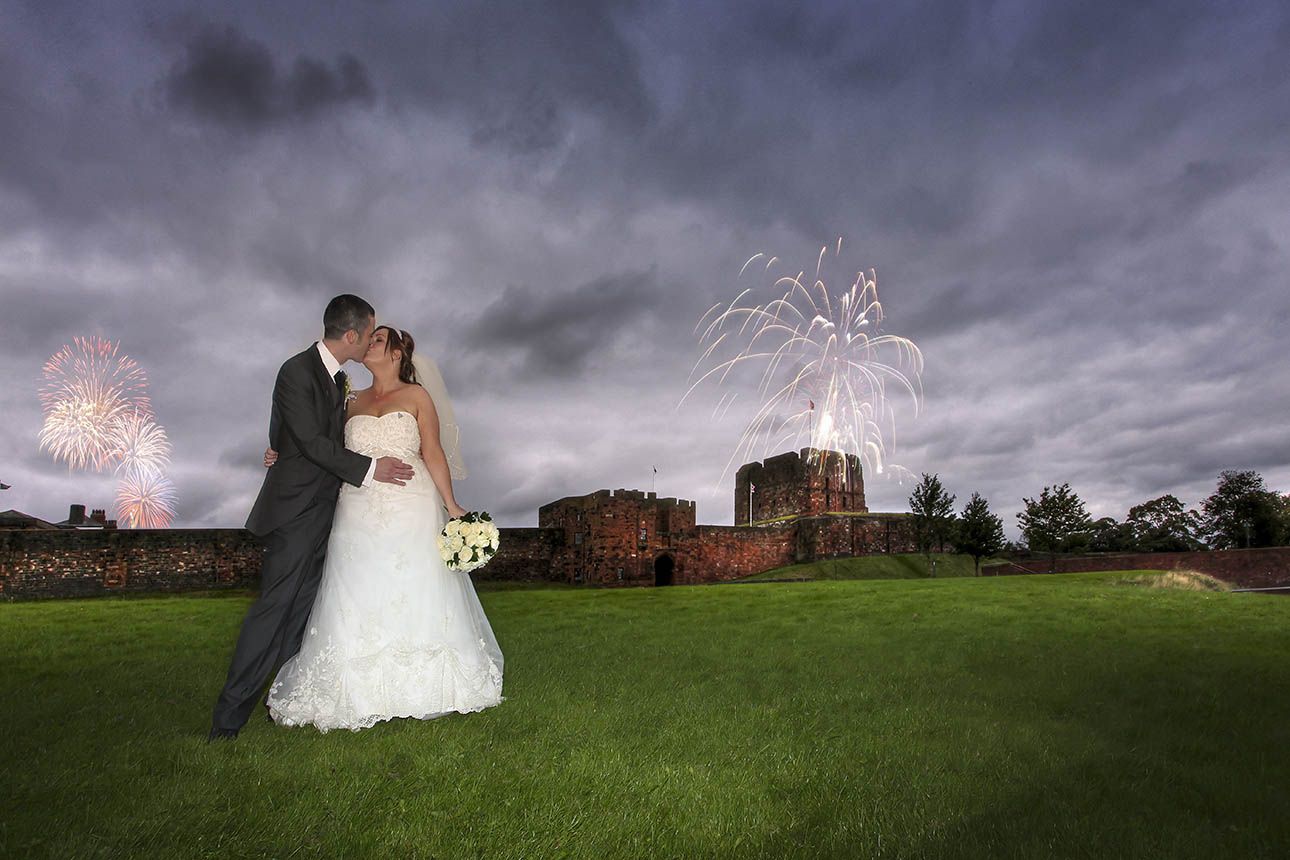 A couple embraces in front of Carlisle Castle at dusk while fireworks light up a dramatic cumbrian sky.