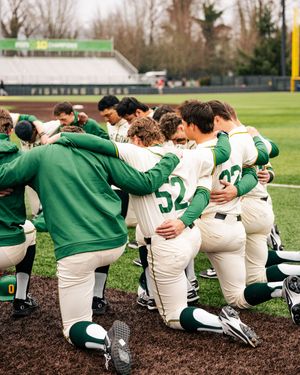 Baseball team members in Oregon uniforms kneel together on the field's warning track.