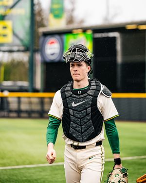 Baseball catcher in white uniform and black protective gear stands ready in position on the field.