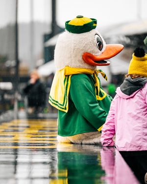 University of Oregon mascot in green and yellow attire watches a rainy game from the sidelines.