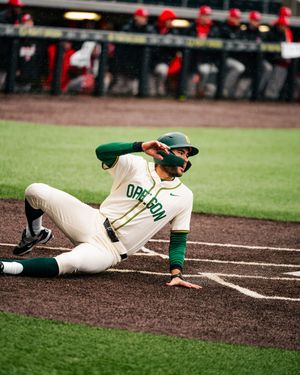 Baseball player in green and white uniform slides across home plate during game action sequence.