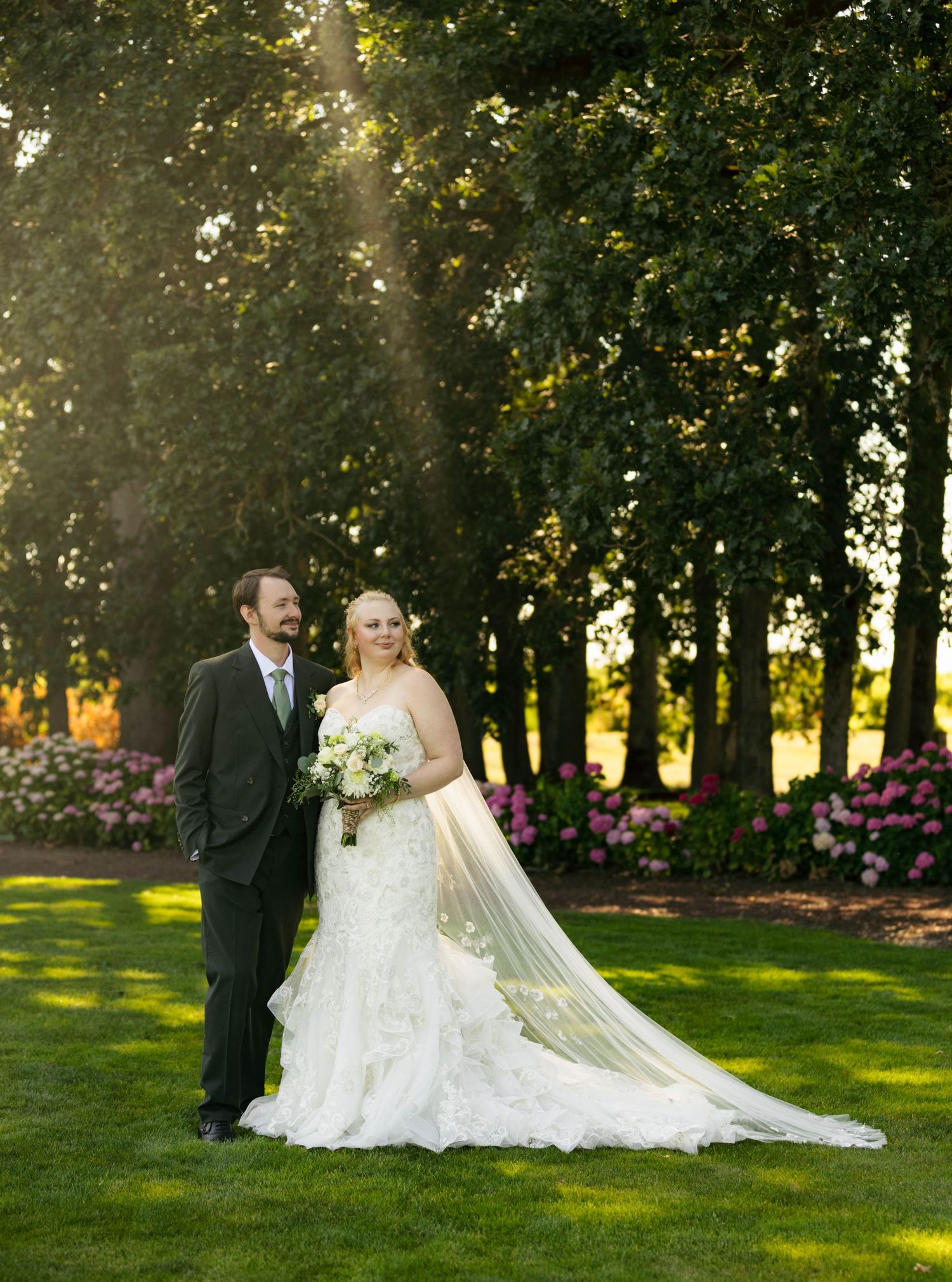 A bride and groom pose together in front of lush green hedges in an elegant garden setting.