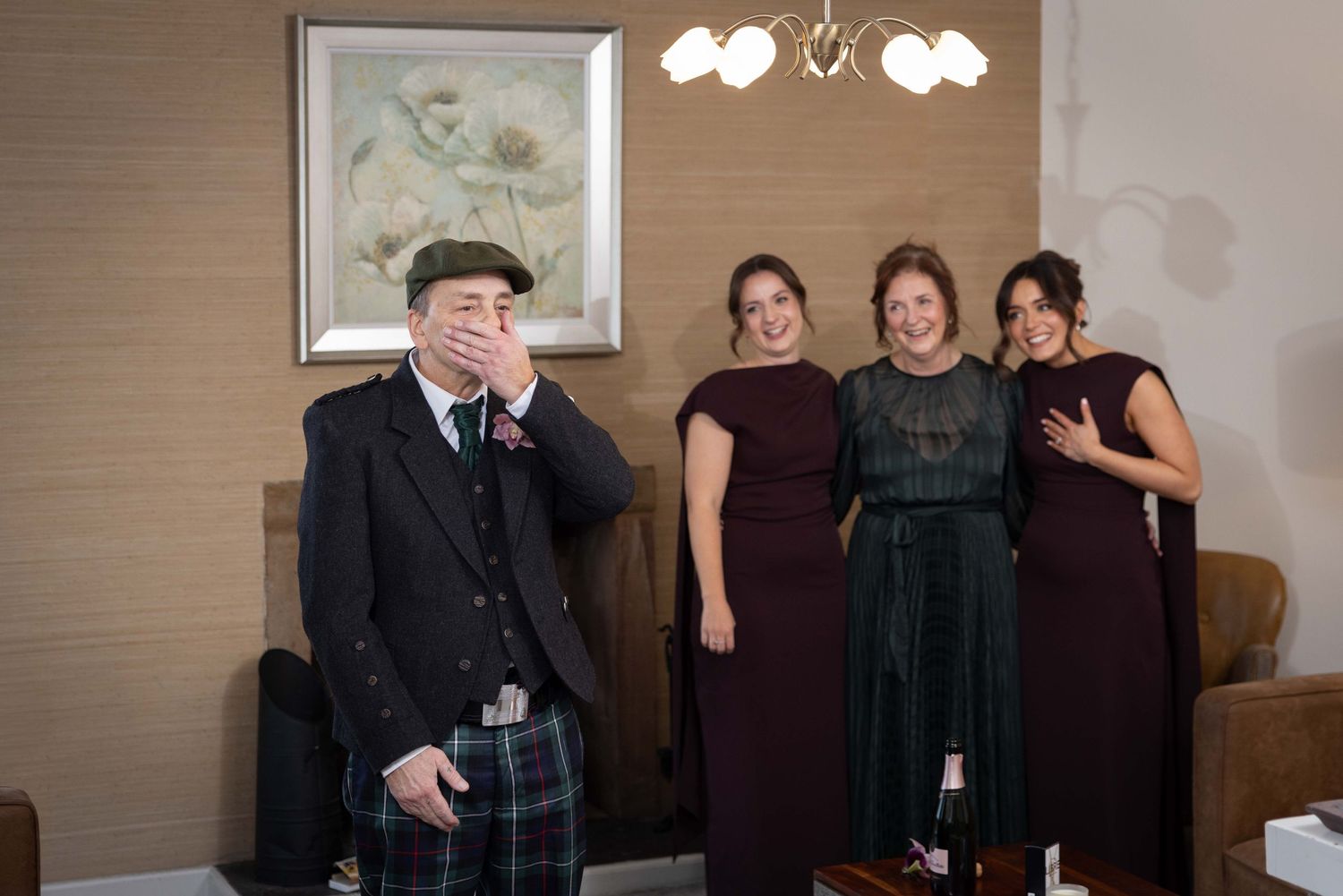 A person in traditional Scottish kilt and cap stands with three others wearing formal dresses at an indoor event.