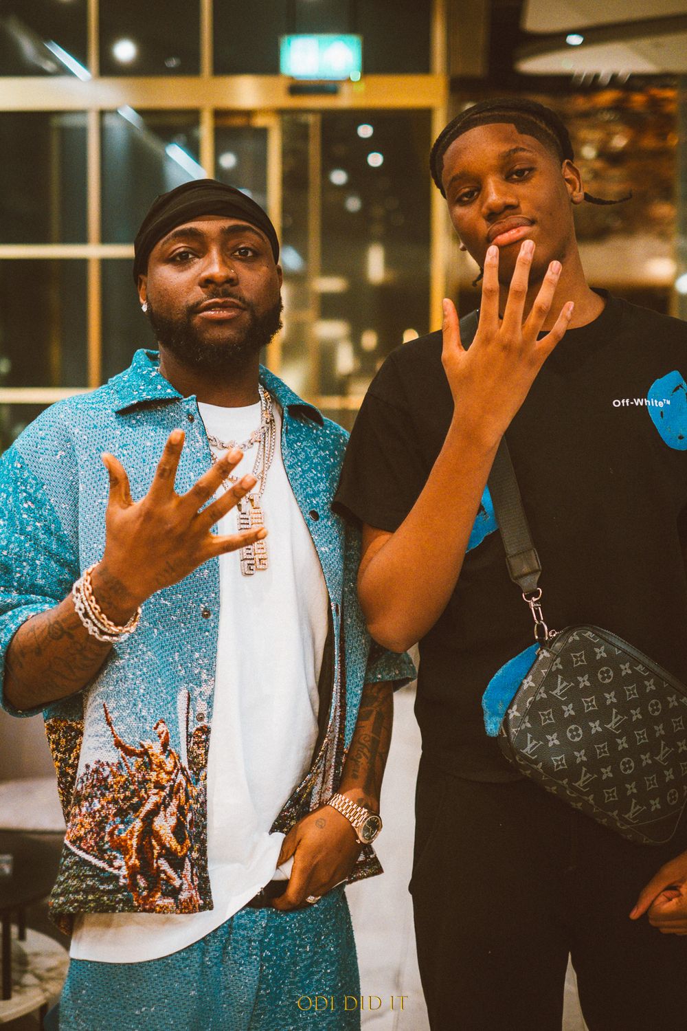 Two people posing together indoors wearing casual attire and showing hand gestures in a nighttime setting.