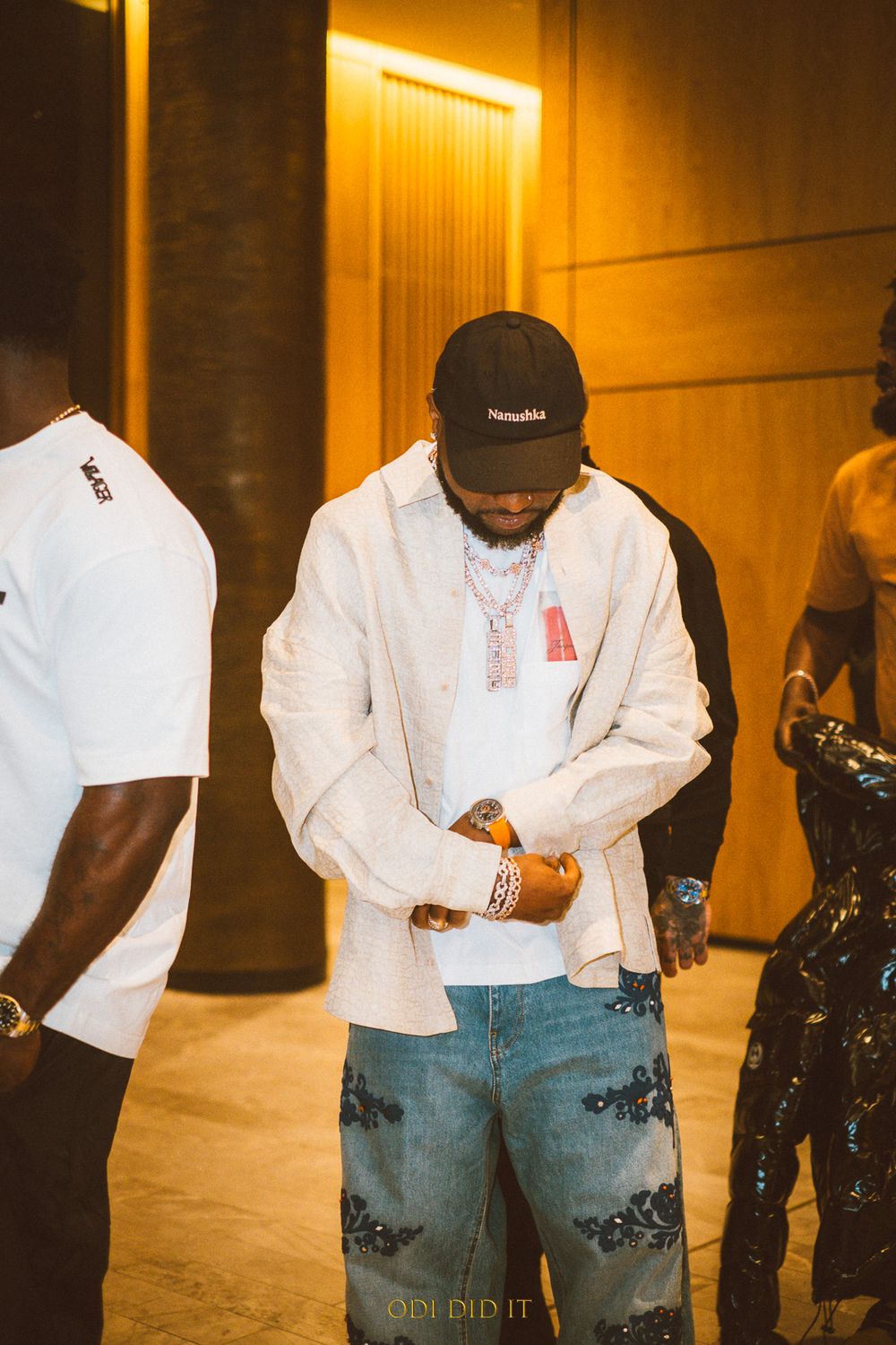 Someone wearing a white button-up shirt, black cap, and distressed jeans stands in a warmly lit hallway.