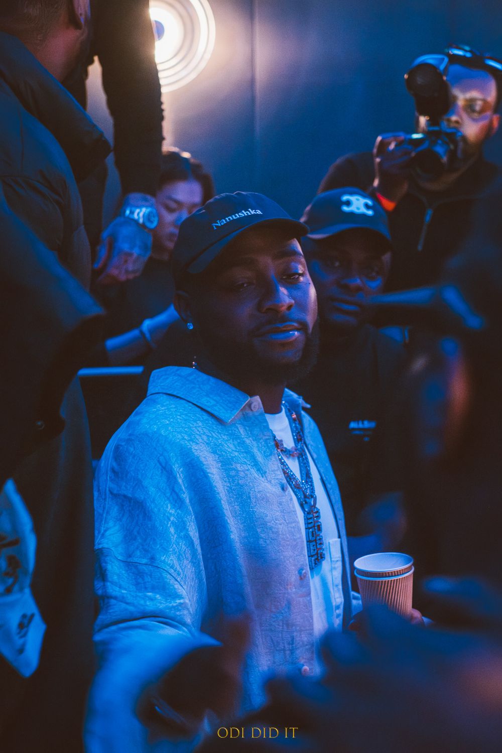 Person in a light blue hoodie smiles while holding a drink cup in a dimly lit venue with blue lighting.