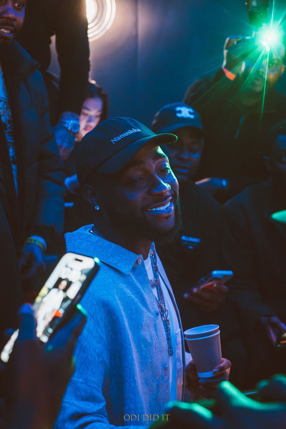 Person in a blue hoodie smiling at a nighttime event with green lights and camera flashes in the background.