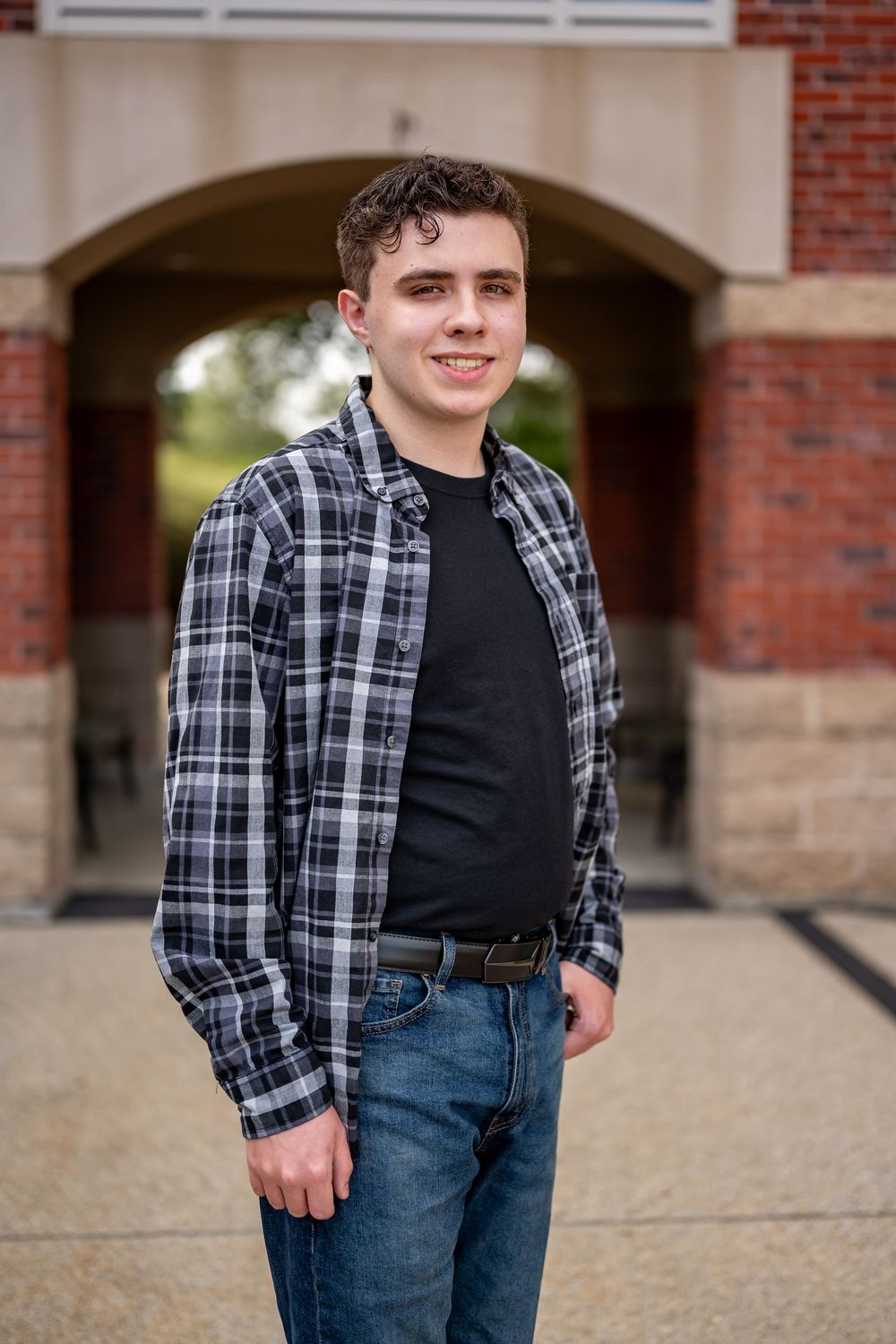 A person wearing a black and white plaid flannel shirt and jeans poses against a brick building with arched doorways.