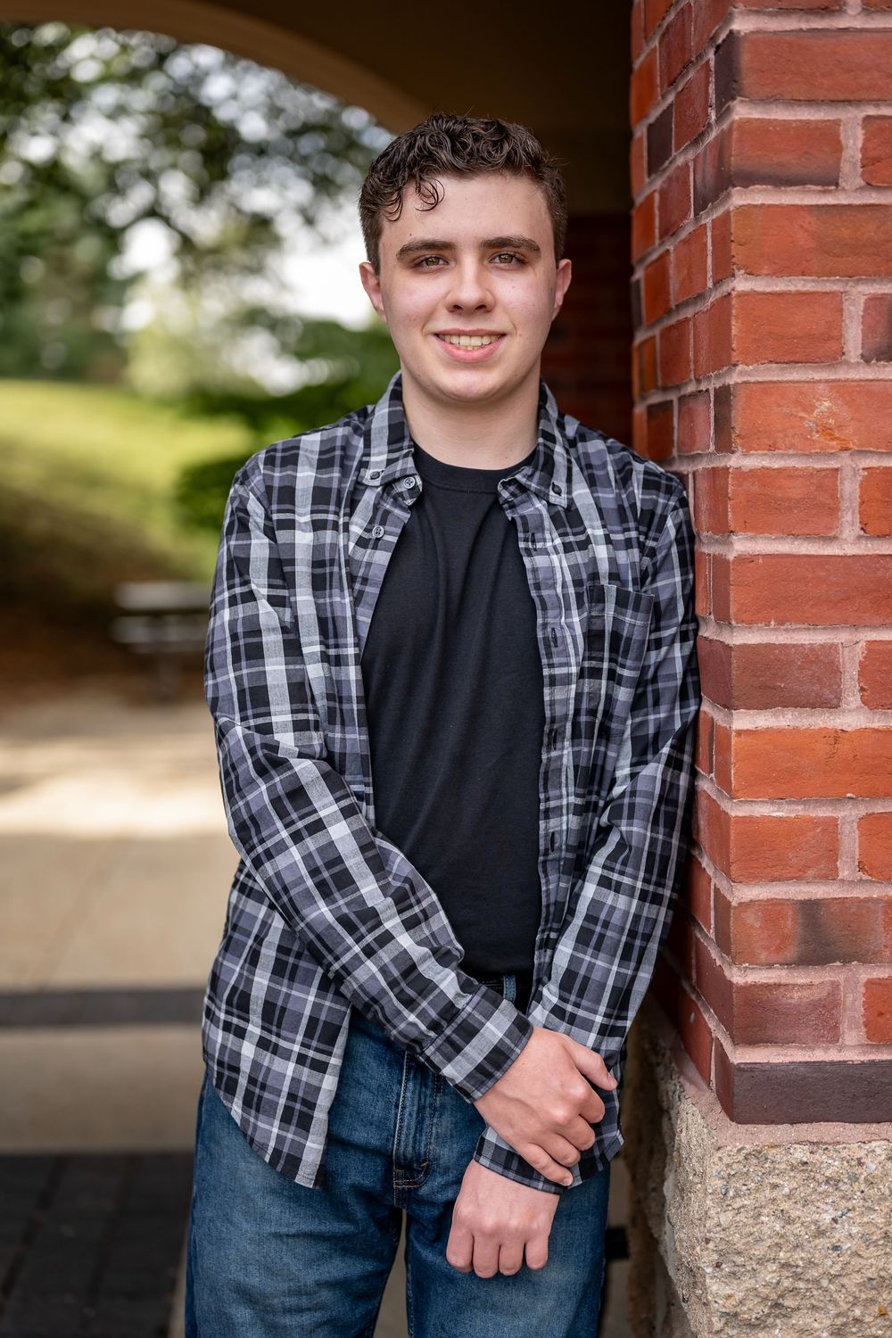 Young person in plaid shirt and jeans poses casually against red brick wall outdoors.