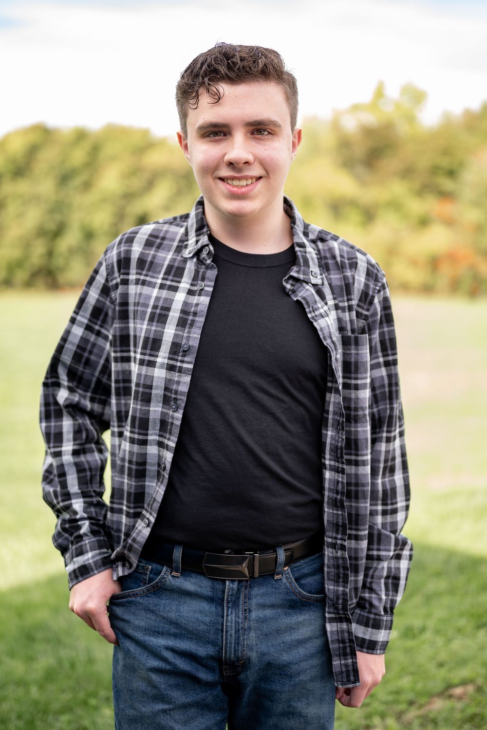 A casual outdoor portrait featuring someone in a plaid button-down shirt and dark jeans in a grassy field.