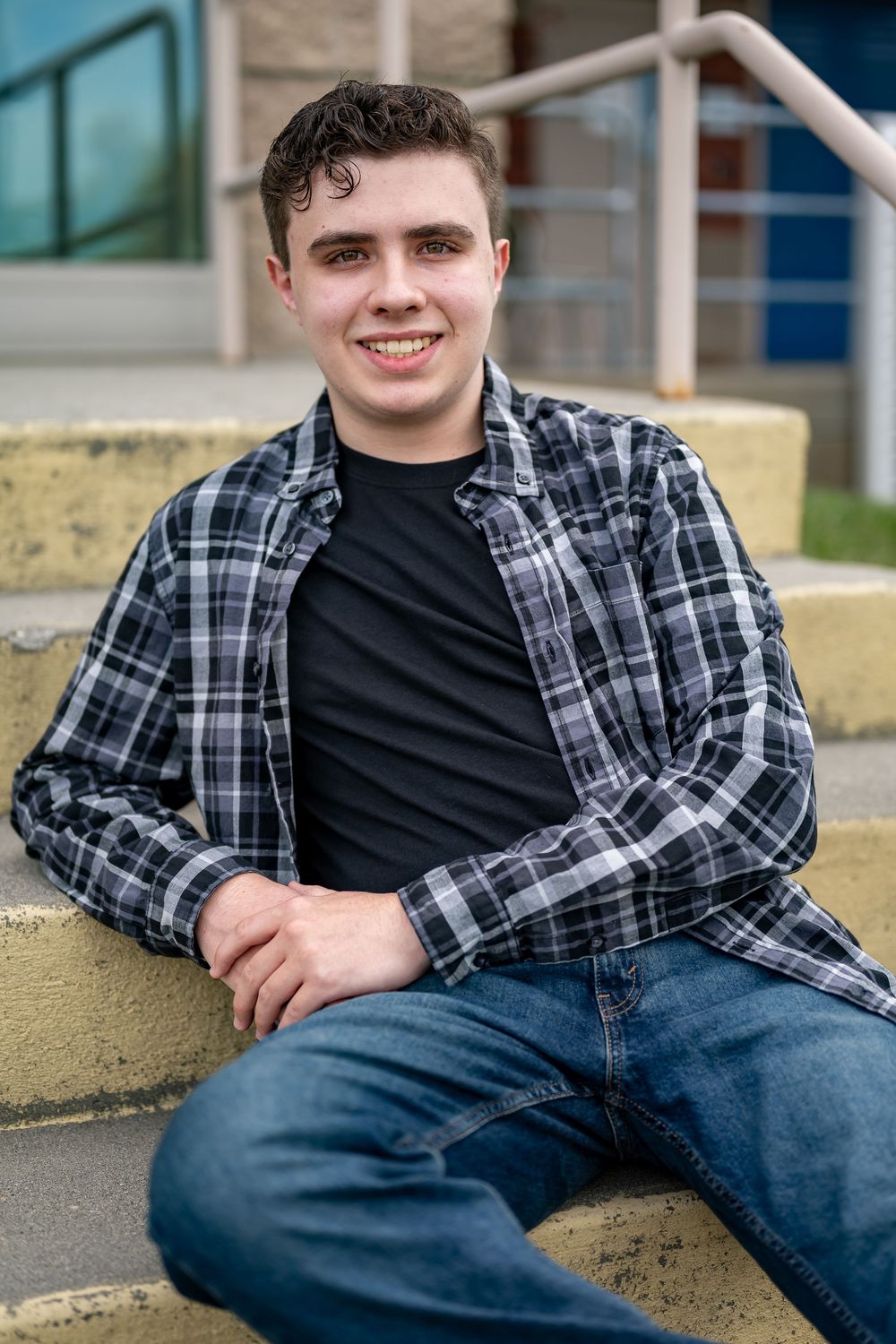 A relaxed portrait of someone sitting on concrete steps wearing a plaid shirt and blue jeans.