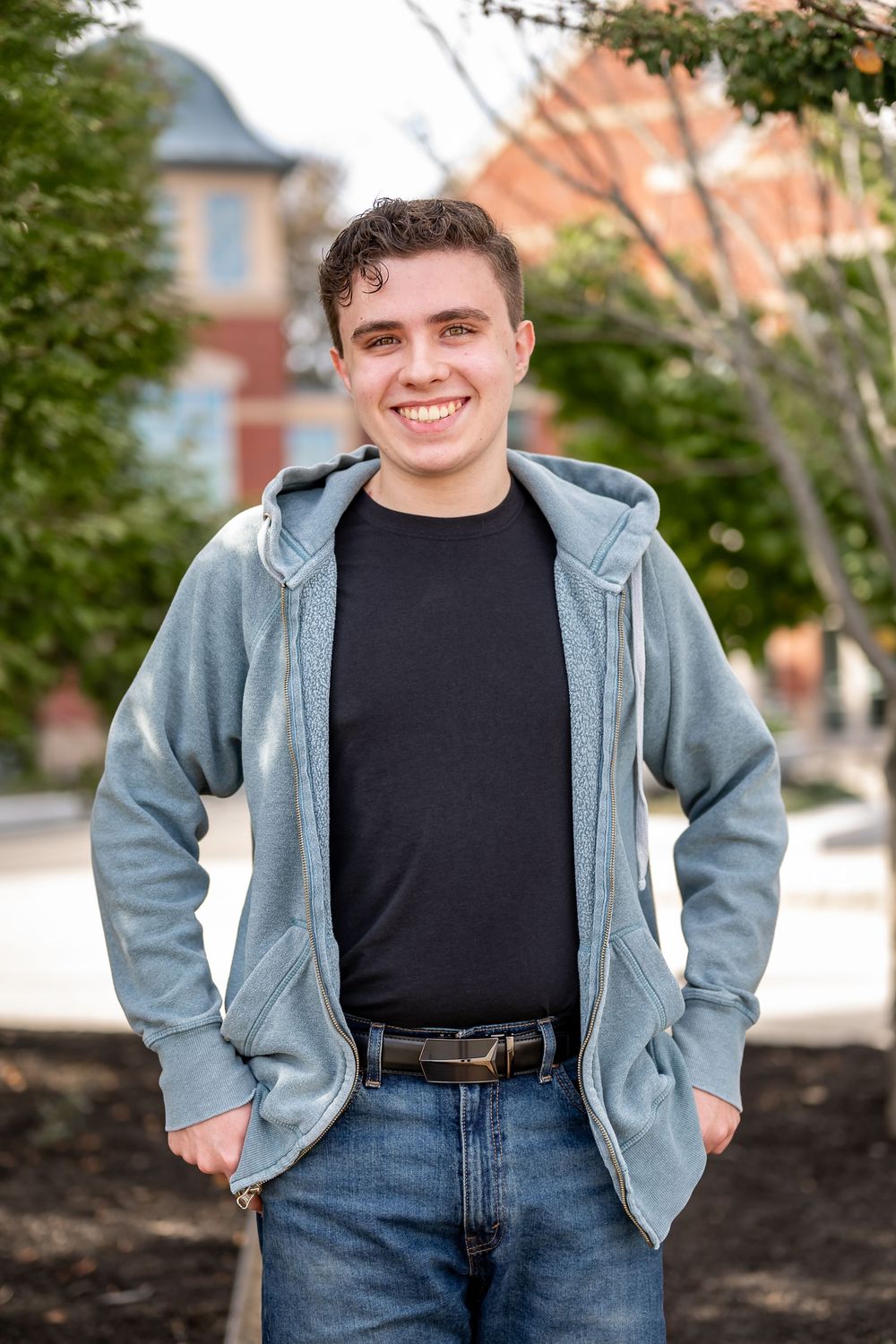 Person wearing a light blue zip-up hoodie and jeans poses outdoors against a backdrop of autumn trees and buildings.