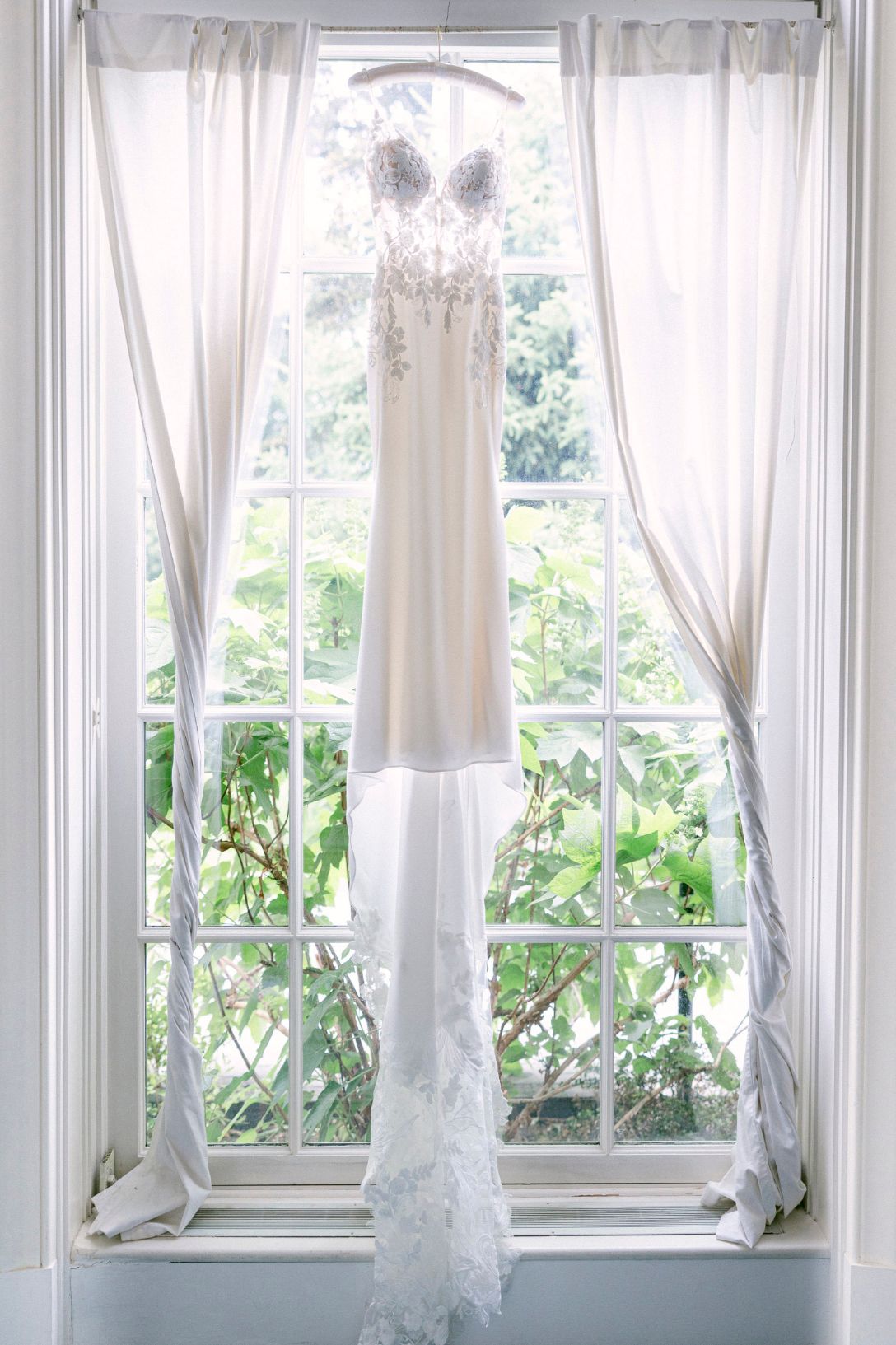 White sheer curtains billow gently in front of a row of bright windows with green plants visible outside.