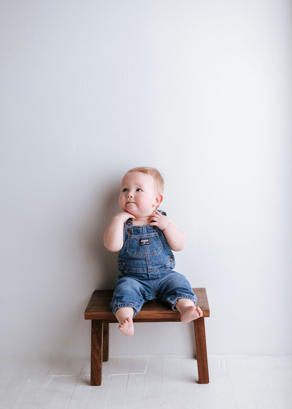 Baby in denim overalls sitting on wooden bench against white background.