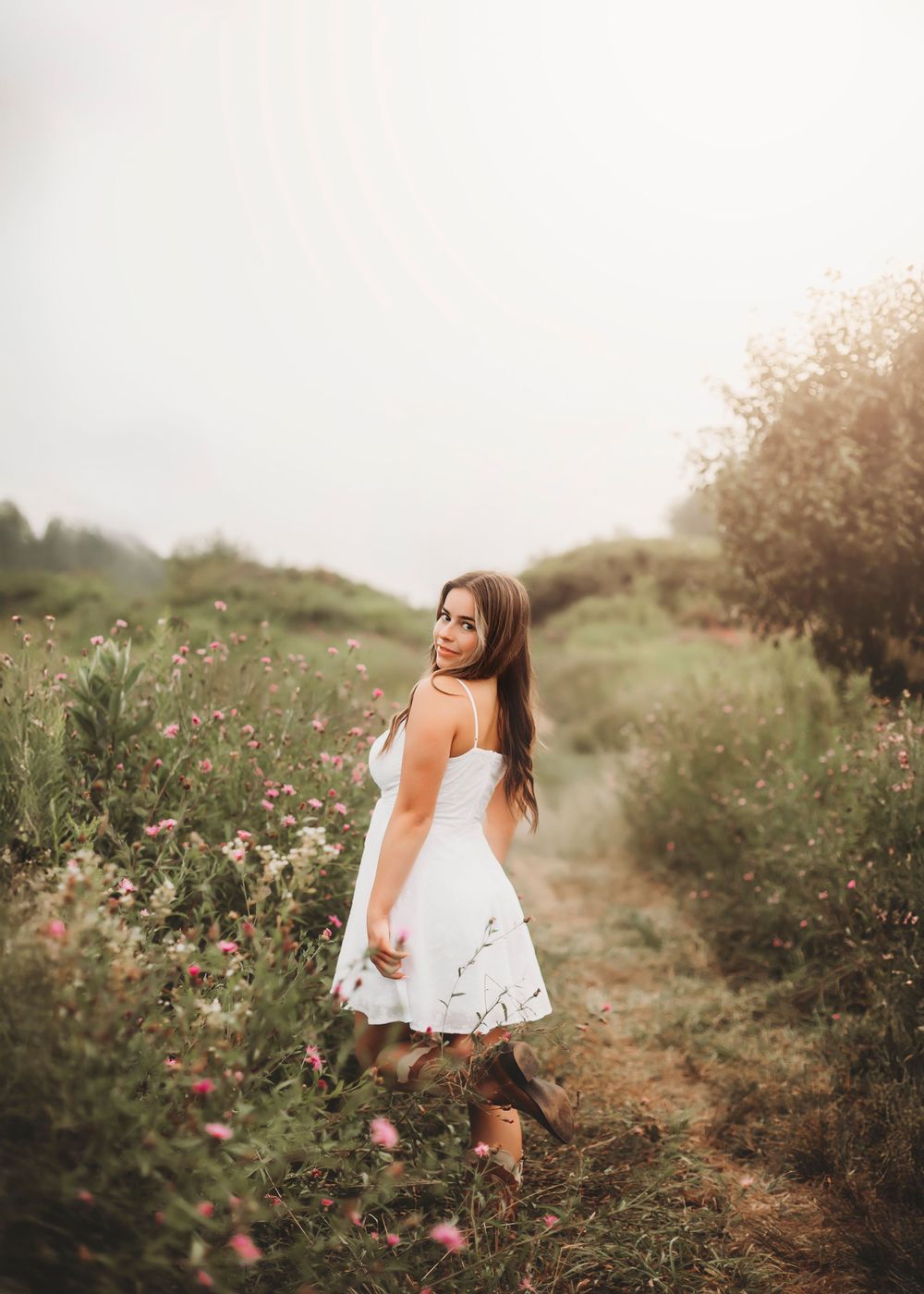 A woman in a white sundress twirls on a dirt path surrounded by wildflowers and greenery at sunset.