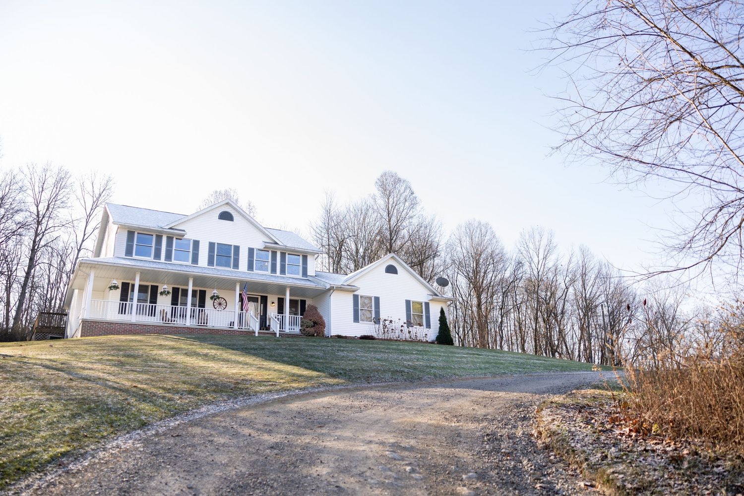 A white colonial style house sits on a hilltop surrounded by bare winter trees and a winding driveway.